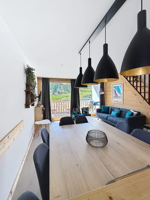 Modern alpine apartment dining area with black pendant lights, wooden table, mountain views, and blue sofa in Montgenèvre