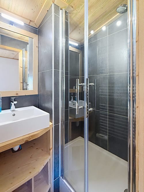Modern bathroom with walk-in shower, white sink, wooden vanity and dark tile walls at Les Tanières de Montgenèvre
