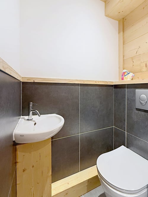 Modern bathroom in Les Tanières duplex with dark tile walls, white corner sink, toilet, and wooden beam ceiling in