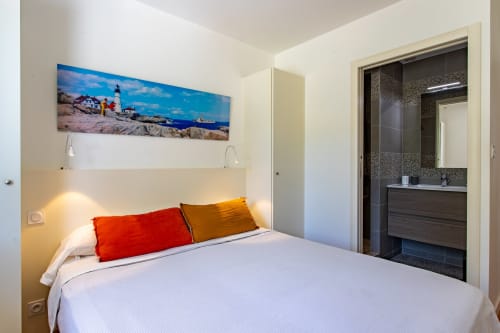 Chambre moderne de villa avec lit blanc, coussins rouges et jaunes, art phare côtier, salle de bains attenante sombre