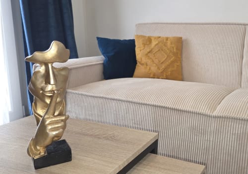 Busto escultórico dorado sobre mesa de madera frente al sofá a rayas beige con cojines azules y dorados en el apartamento Le Contemporain en NN