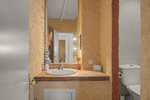 Baño rústico con paredes de terracota, lavabo blanco, espejo, inodoro y mostrador de madera en Can Gat Vell B&B en Girona