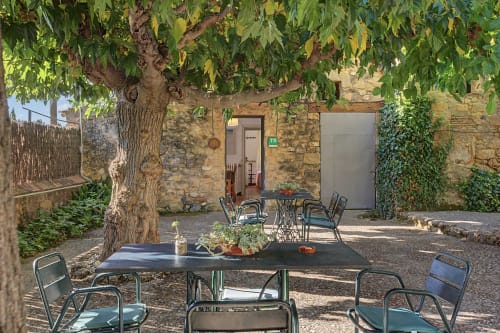 Patio exterior sombreado de Can Gat Vell en Girona con asientos, árbol grande, edificio de piedra y plantas en macetas