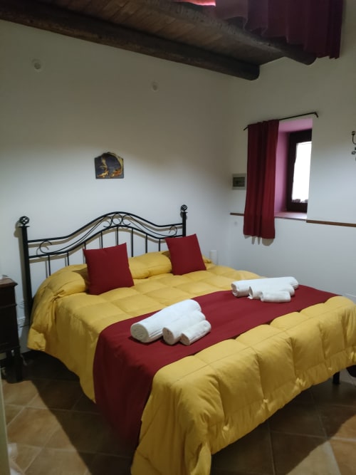 Camera da letto con biancheria gialla e bordeaux, cuscini decorativi, asciugamani arrotolati, letto in ferro battuto, soffitto in legno a vista