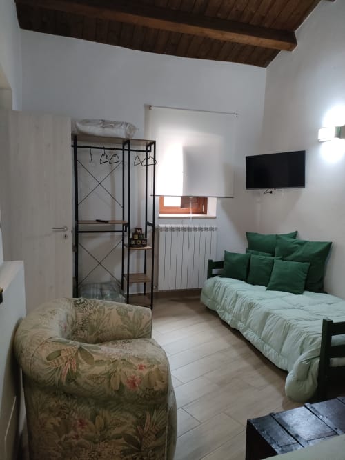 Camera da letto luminosa con cuscini verdi, poltrona decorata, TV schermo piatto e soffitto con travi in legno presso Case Vacanze