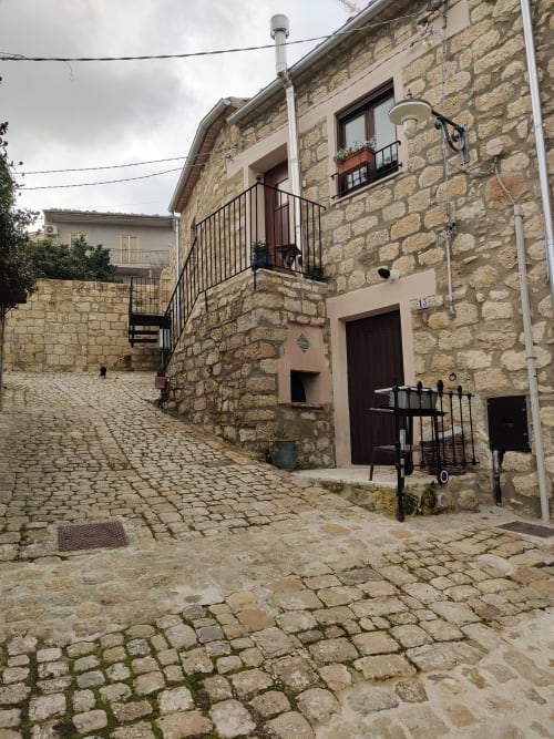 Ingresso in pietra di edificio storico con terrazza affacciata sulla piazza del borgo di Petralia Soprana, Case Vacanze Borgo i Stritti