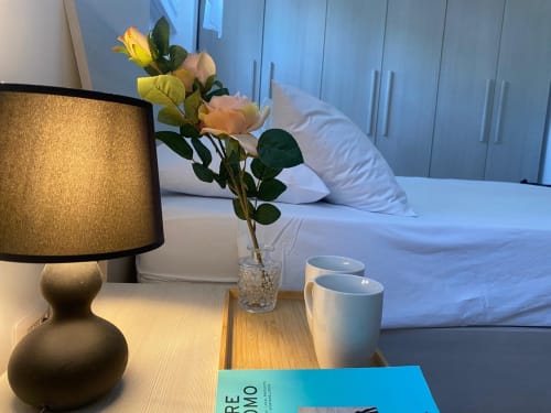 Comodino della camera da letto dell'appartamento Le case di Angy con lampada, rose gialle, bicchiere d'acqua e tazze di caffè con vista sul letto