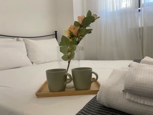 Dettaglio della camera da letto presso Le case di Angy: due tazze salvia, rose fresche in vaso di vetro, vassoio in legno e biancheria bianca sul letto