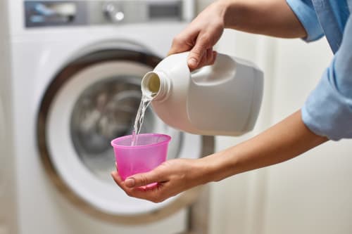 Personne versant un produit de lessive dans une tasse rose près d'une machine à laver à La Grange Aux Etangs
