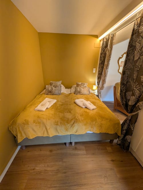 Cozy cottage bedroom at Bi d'Grosseltere in Kirrwiller with mustard yellow walls, queen bed, patterned curtains, and wooden