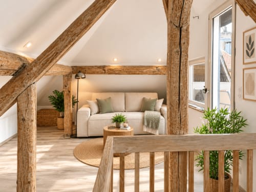 Loft à Metz avec poutres en bois apparent, canapé crème, meubles bois et vue sur la ville depuis la porte-balcon