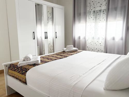 Spacious bedroom at DakarByDays apartment with white bedding, decorative runner, twin mirrors, white wardrobe, and sheer
