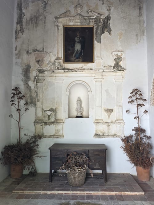 Capilla rústica en Hacienda Las Lumbreras con columnas ornamentadas, retrato, nicho con estatua y flores secas en macetas de barro