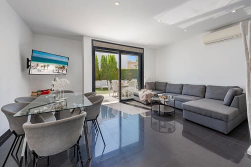 Modern villa living room in Alcudia with gray sectional sofa, dining area, TV, and terrace access with garden views