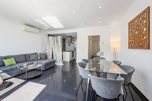 Modern villa living area in Alcudia with dining table, gray sofa, open kitchen, and decorative wood panel art