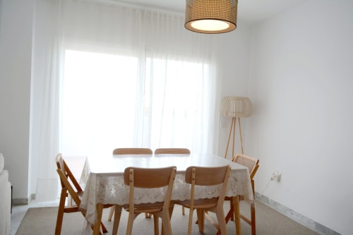 Comedor luminoso en apartamento Royal Banus, Marbella, con sillas de madera, mesa y cortinas translúcidas con vistas a la costa