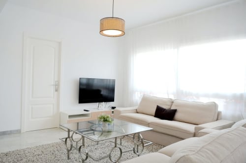 Moderno salón con sofá beige, televisor de pantalla plana, mesa de centro de cristal y lámpara colgante en Royal Banus