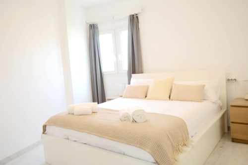 Dormitorio moderno y luminoso en Royal Banus, Marbella, con cama queen, ropa de cama crema, cortinas grises y apartamento costero