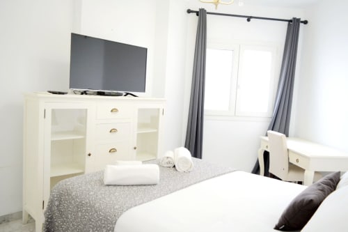 Modern bedroom at Royal Banus Marbella with white furniture, grey patterned bedding, TV, and grey curtains by balcony door
