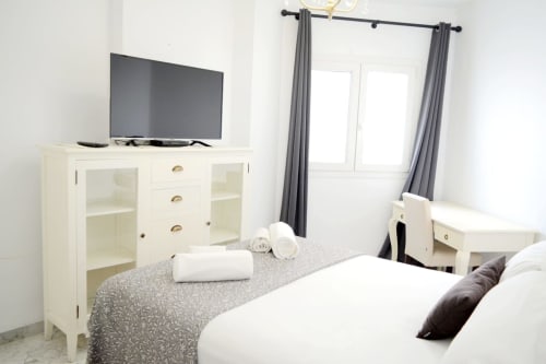 Modern bedroom at Royal Banus Marbella with white bed, cream dresser, TV, and gray curtains overlooking balcony