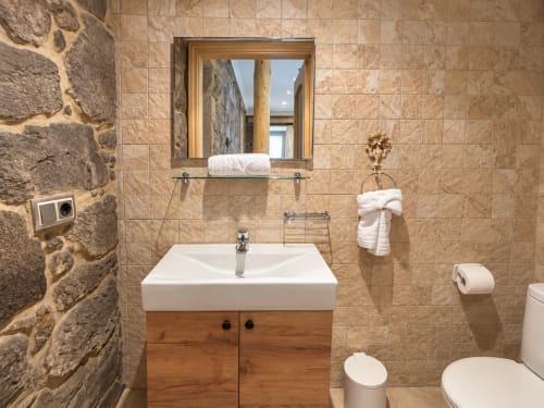 Baño moderno en apartamento de Baiona con paredes de piedra, lavabo de madera, espejo, toallas blancas y luz natural abundante
