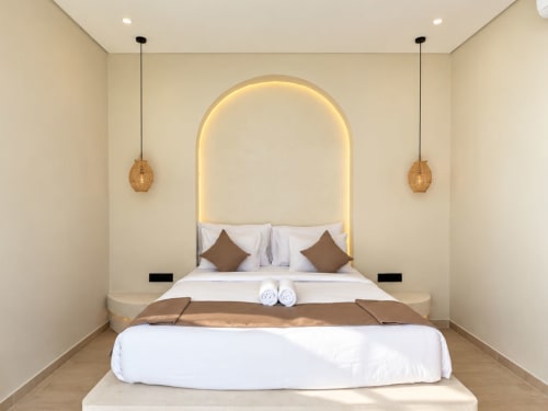 Dormitorio de villa moderna en Oasis Dune Villas con cabecero de neón arqueado, almohadas marrones, lámparas colgantes y ropa de cama blanca