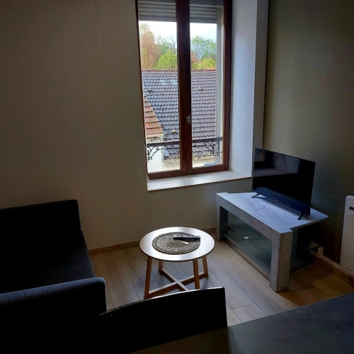 Élégant séjour moderne aux Hortensias avec téléviseur, table ronde, chaises noires et portes-fenêtres donnant sur les toits en tuiles