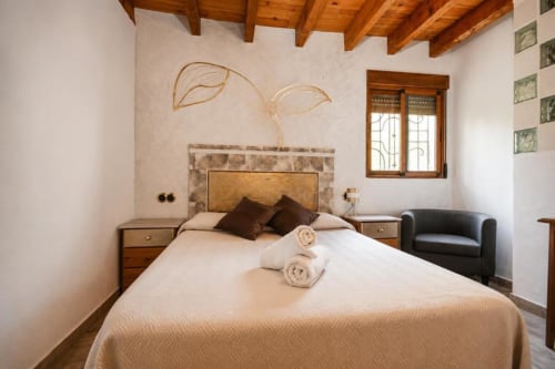 Rustic bedroom with wooden beamed ceiling, king-size bed with brown pillows, leaf artwork above headboard, and black armchair