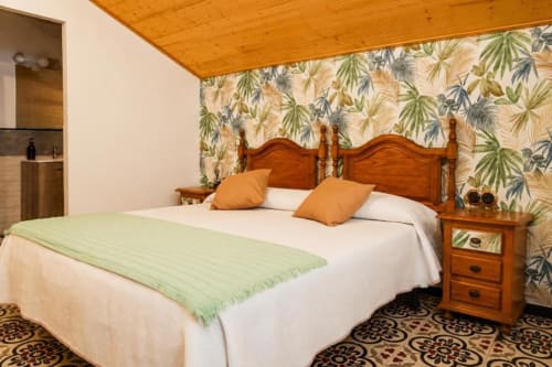 Cozy triple room at Serranía hotel in Chulilla with wooden bed, orange pillows, tropical wallpaper, and garden views