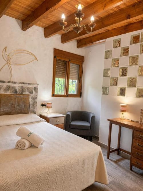 Rustic bedroom with exposed wood beams, cream bedding, dark armchair, and decorative wall tiles at Serranía hotel in Chulilla
