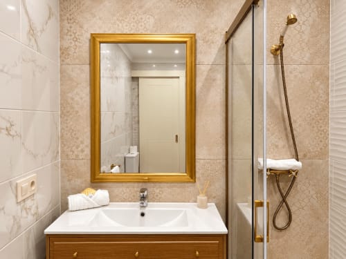Baño moderno en apartamento Holibai, Baiona con espejo de marco dorado, azulejos de mármol, ducha tipo lluvia y tocador de madera