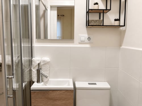 Baño moderno en apartamento Holibai de Baiona con paredes de azulejos blancos, espejo iluminado, tocador de madera, inodoro y ducha