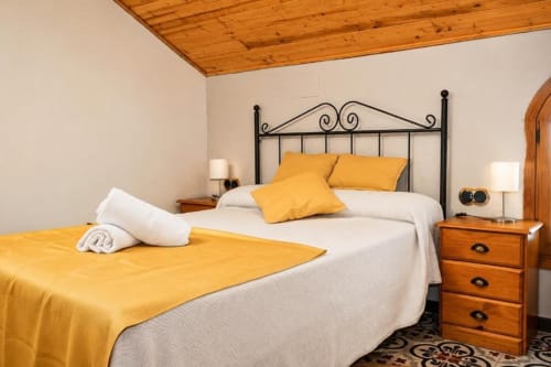 Bright bedroom with yellow accents, wrought iron bed, wooden nightstands, and wooden ceiling in Chulilla hotel
