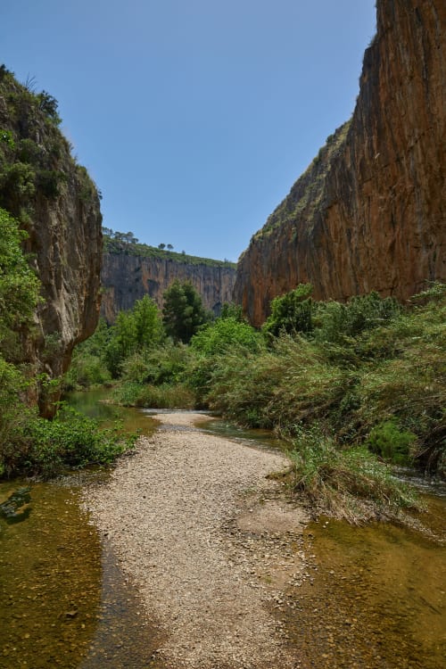 Narrow canyon gorge with towering rock cliffs, shallow river, and lush vegetation in Chulilla, Spain