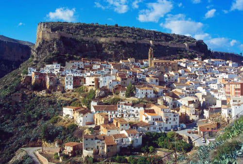 Chulilla village nestled below dramatic cliff formations with colorful houses cascading down the hillside under blue sky