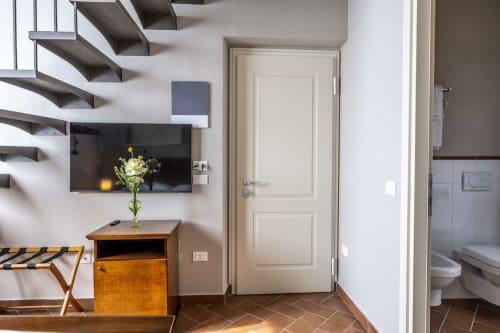 Camera moderna al Corte Baruzzo Relais con scala sospesa, TV a schermo piatto, comodino in legno e porte color crema che si aprono su