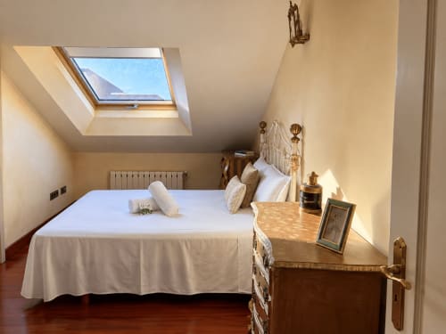 Luminoso dormitorio abuhardillado en Baiona con tragaluz, cama de hierro blanco, muebles de madera y vista al mar desde la ventana