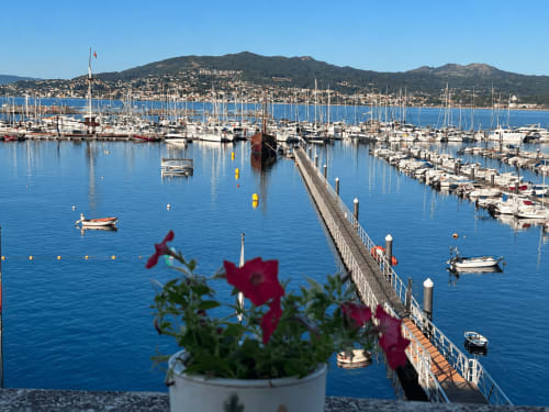 Marina de Baiona, España con veleros, flores rojas, casas en la ladera y aguas azules cristalinas—vista desde terraza junto al mar