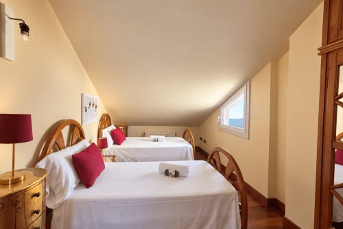 Luminoso dormitorio abuhardillado en Baiona con dos camas, almohadas burdeos, muebles de madera, ventana con vista al mar e iluminación cálida