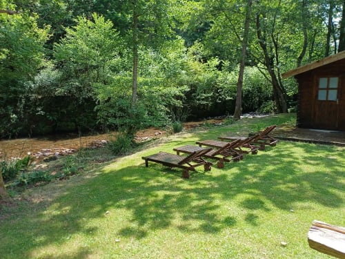 Wooden lounge chairs on green lawn beside a stream at Le gîte des Mousquetaires cottage, surrounded by forest trees in Lanne
