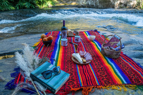 Piquenique colorido sobre manta tecida à beira do rio em The Ranch Project.Co, Gonçalves Brasil, com vinho, petiscos e montanha