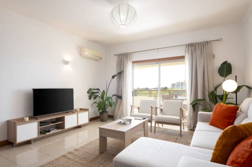 Bright modern apartment living room at RB Algarve Vilamoura with sofa, TV, balcony view, plants and contemporary furnishings.