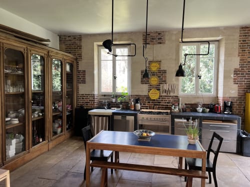 Cuisine très bien équipée, charme des matériaux et de l'espace aux murs de brique, luminaires noirs suspendus, table en bois et vue sur le jardin aux Anciennes écuries du château