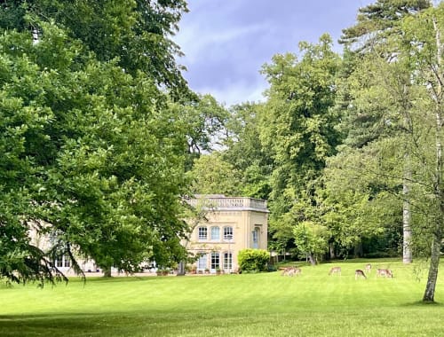 Parc du château avec bâtisse de pierre claire, pelouse impeccable, daims en pâturage et arbres centenaires aux Anciennes Écuries du Château