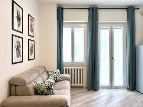 Accogliente salotto milanese con divano beige, opere botaniche, tende turchesi e luminose finestre con radiatore