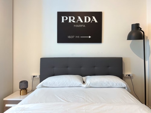 Camera moderna in appartamento milanese con letto rivestito nero, biancheria bianca e cartello Prada Marfa sopra la testata