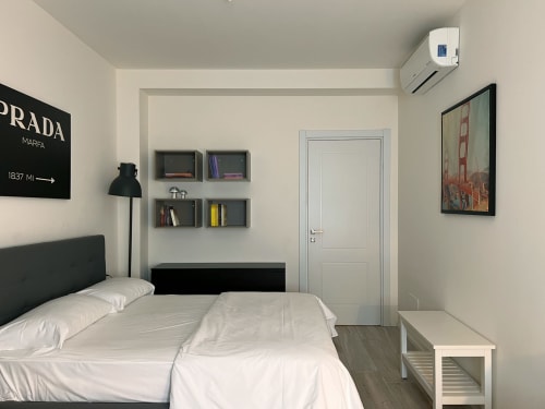 Camera moderna a Nestco Milano con biancheria bianca, testata nera, mensole, poster Prada e arredamento minimalista