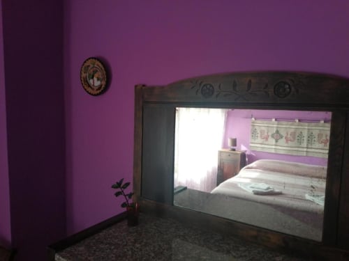 Camera da letto viola presso il B&B Daddanca ad Ales con specchio che riflette il letto, orologio decorativo e pianta su pavimento scuro