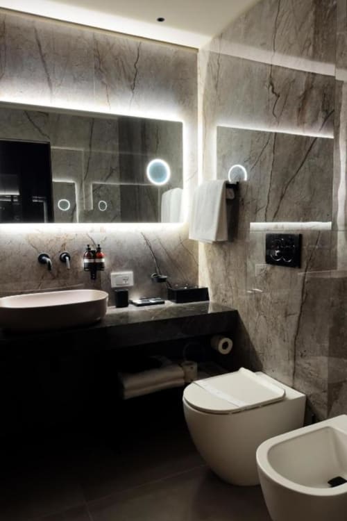 Bagno di lusso moderno con rivestimenti in marmo, specchio illuminato a LED, lavabo bianco a bacinella, bidet e WC da Donna Filomena Luxury