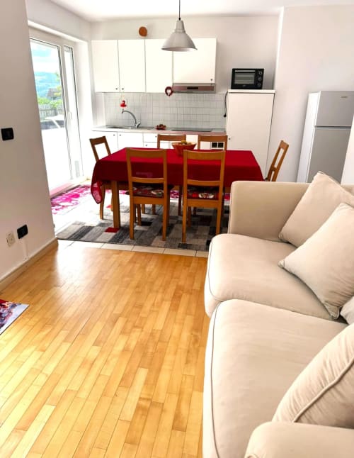 Moderno appartamento a Siusi con divano beige, tavolo rosso da pranzo, pavimenti in legno, cucina e vista sulle montagne dal balcone